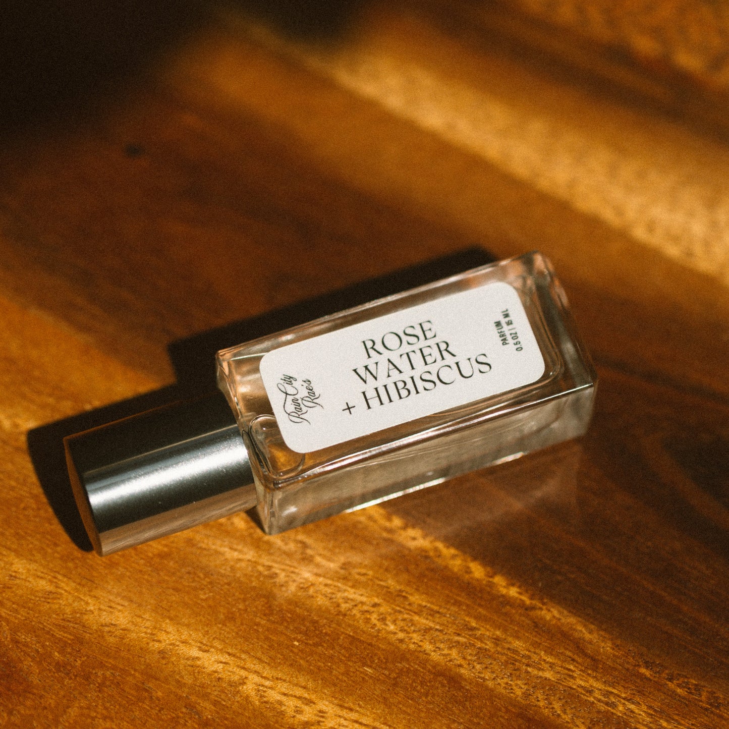 A small clear glass perfume bottle labeled 'ROSE WATER + HIBISCUS' with a silver cap, resting on a warm wooden surface under dramatic golden lighting.