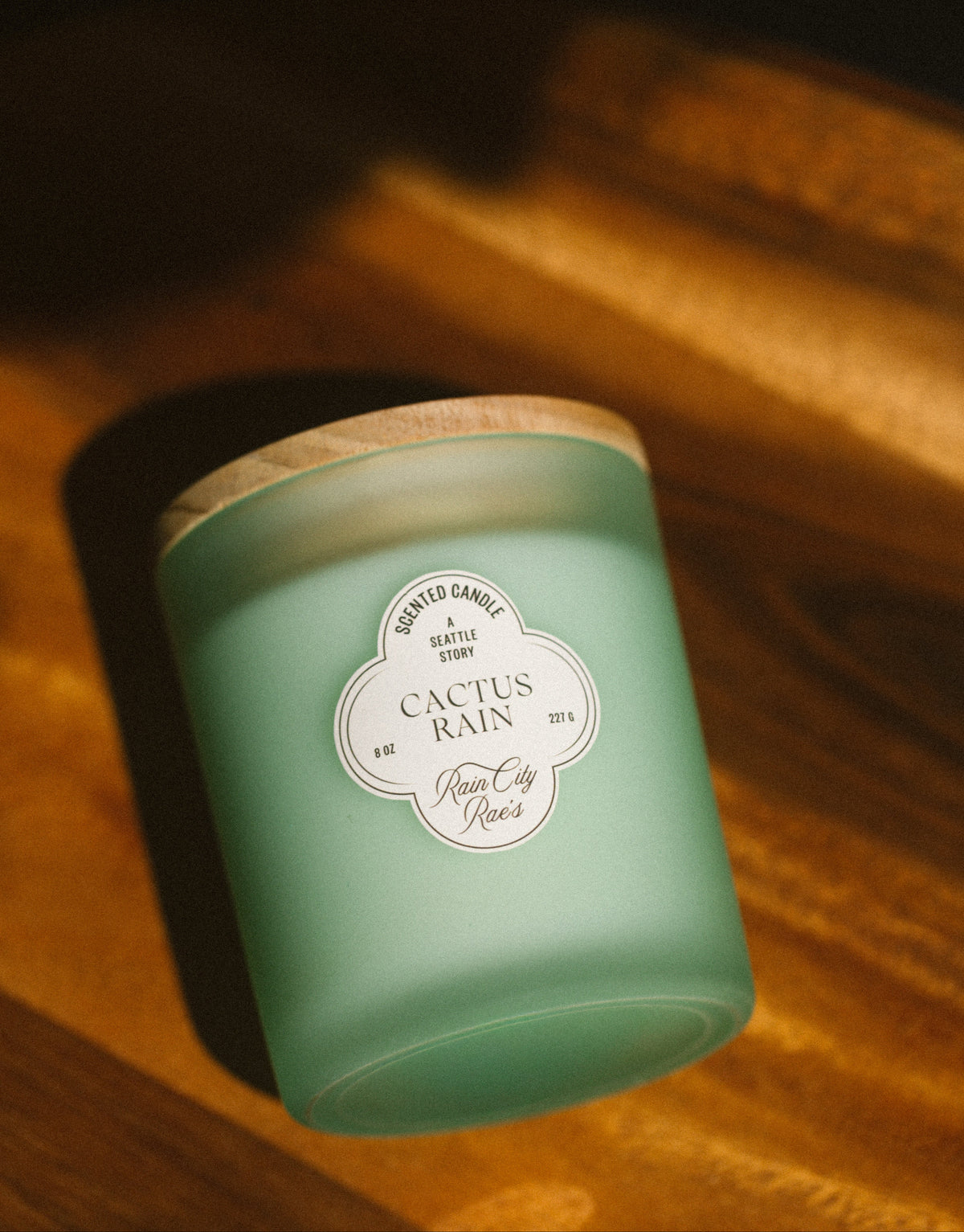 A frosted mint green candle jar with a light wood lid, resting on a warm wooden surface under dramatic golden lighting. The front label reads 'Cactus Rain' By Rain City Rae's