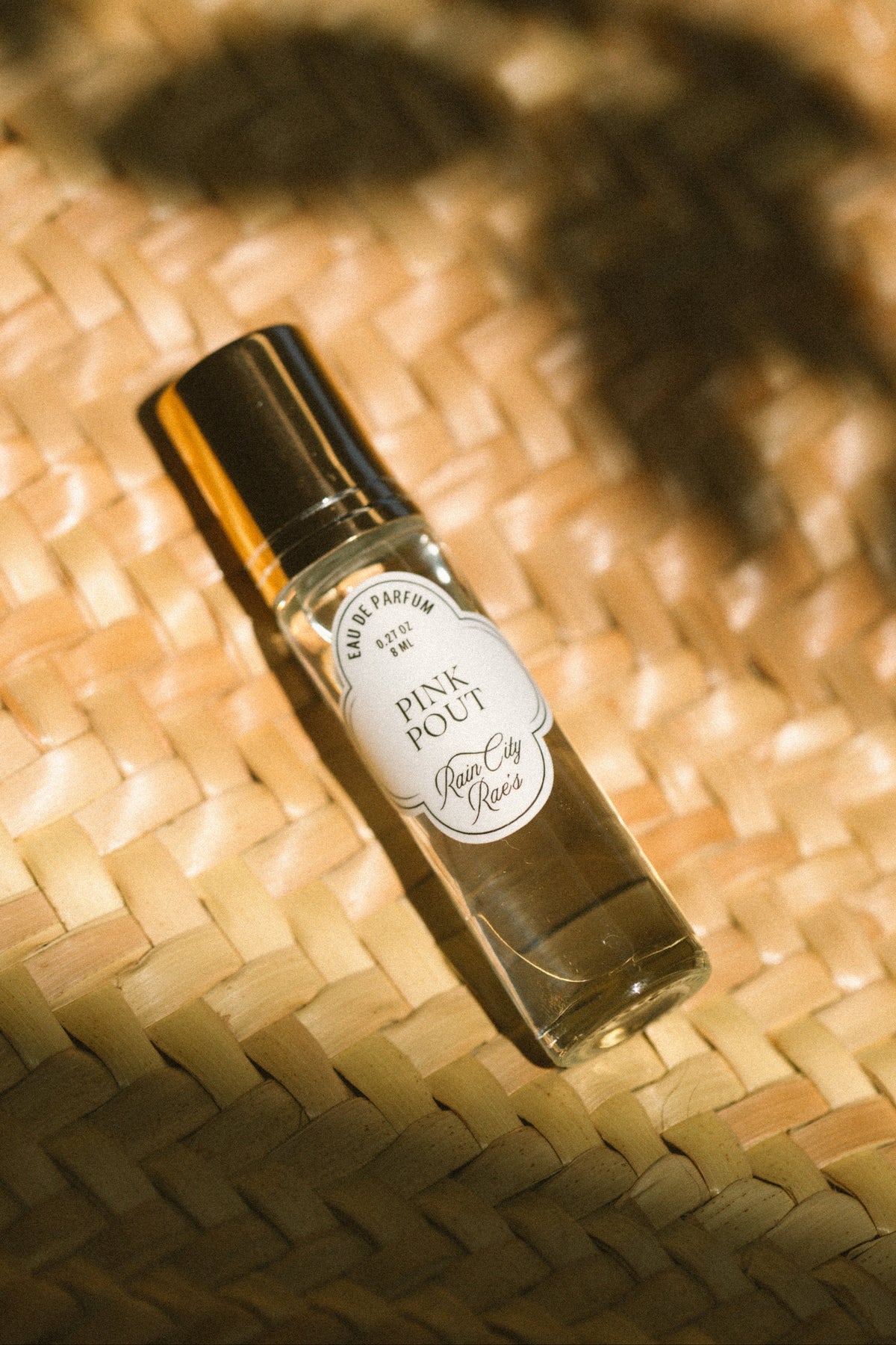 A small glass perfume bottle with a gold cap and a white oval label reading Pink Pout, placed on a woven straw surface with warm sunlight and leaf shadows. By Rain City Rae's