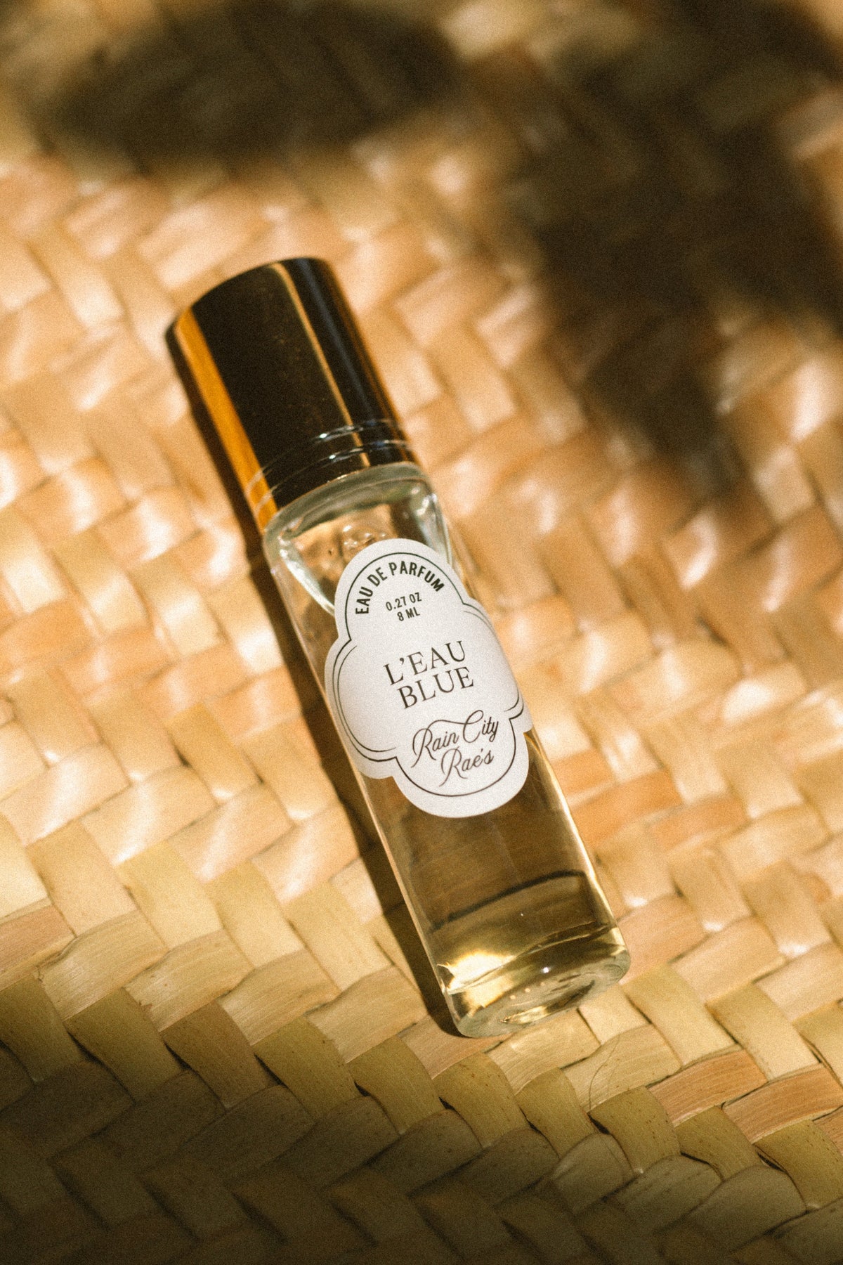 A small clear glass perfume bottle with a gold cap and a white oval label reading 'L'EAU BLUE' By Rain City Rae's, placed on a woven straw surface with warm sunlight and leaf shadows.