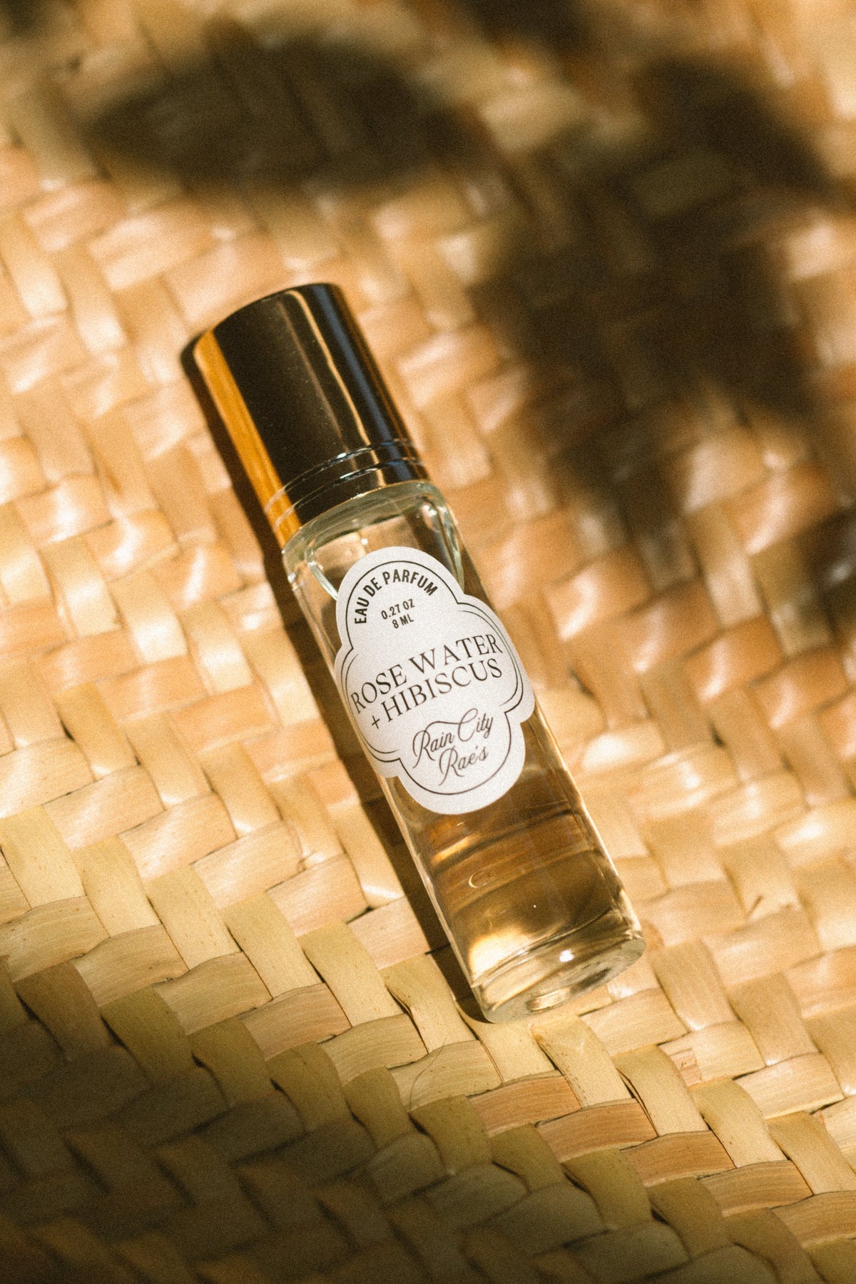 A small clear glass roller bottle perfume with a black cap and a white circular label reading 'Rose Water and Hibiscus' By Rain City Rae's, placed on a woven straw surface with warm sunlight and leaf shadows.