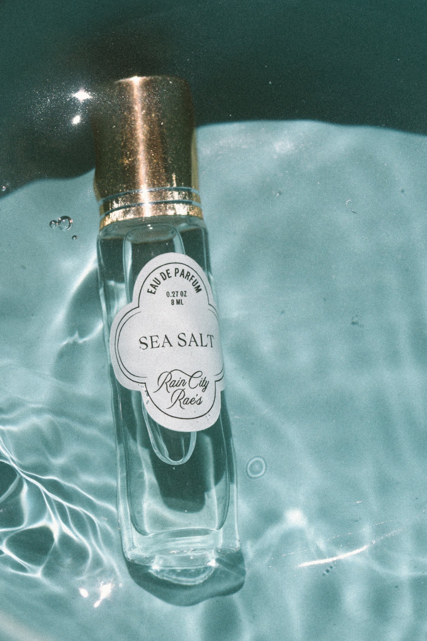 A clear glass perfume roller bottle labeled 'Sea Salt' by Rain City Rae's, featuring a metallic gold cap, resting on rippling blue water.