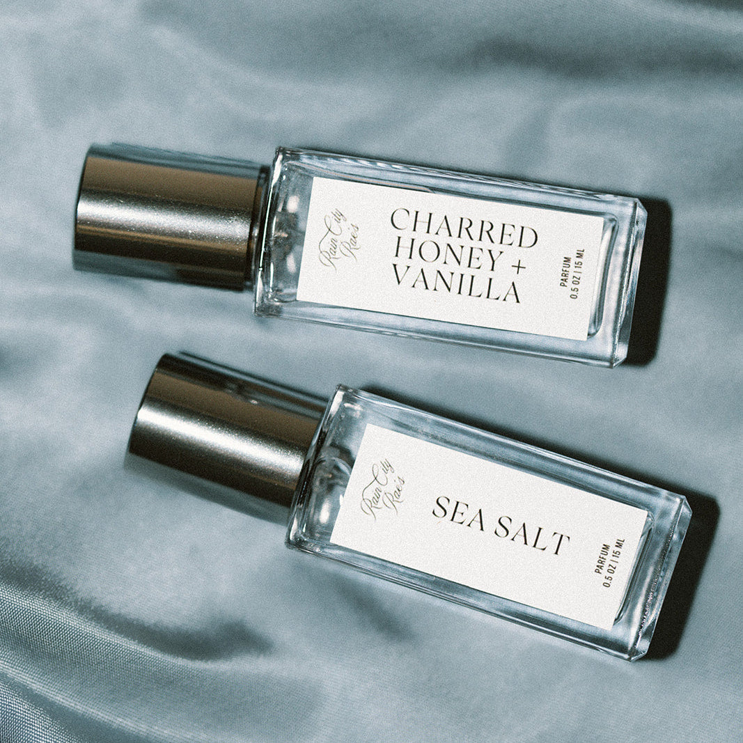 Two perfume bottles labeled 'Charred Honey + Vanilla' and 'Sea Salt' on a textured blue silk surface.