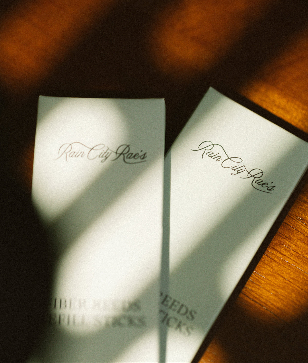 Two diffuser reed refill packs with Rain City Rae's branding on a wooden surface.