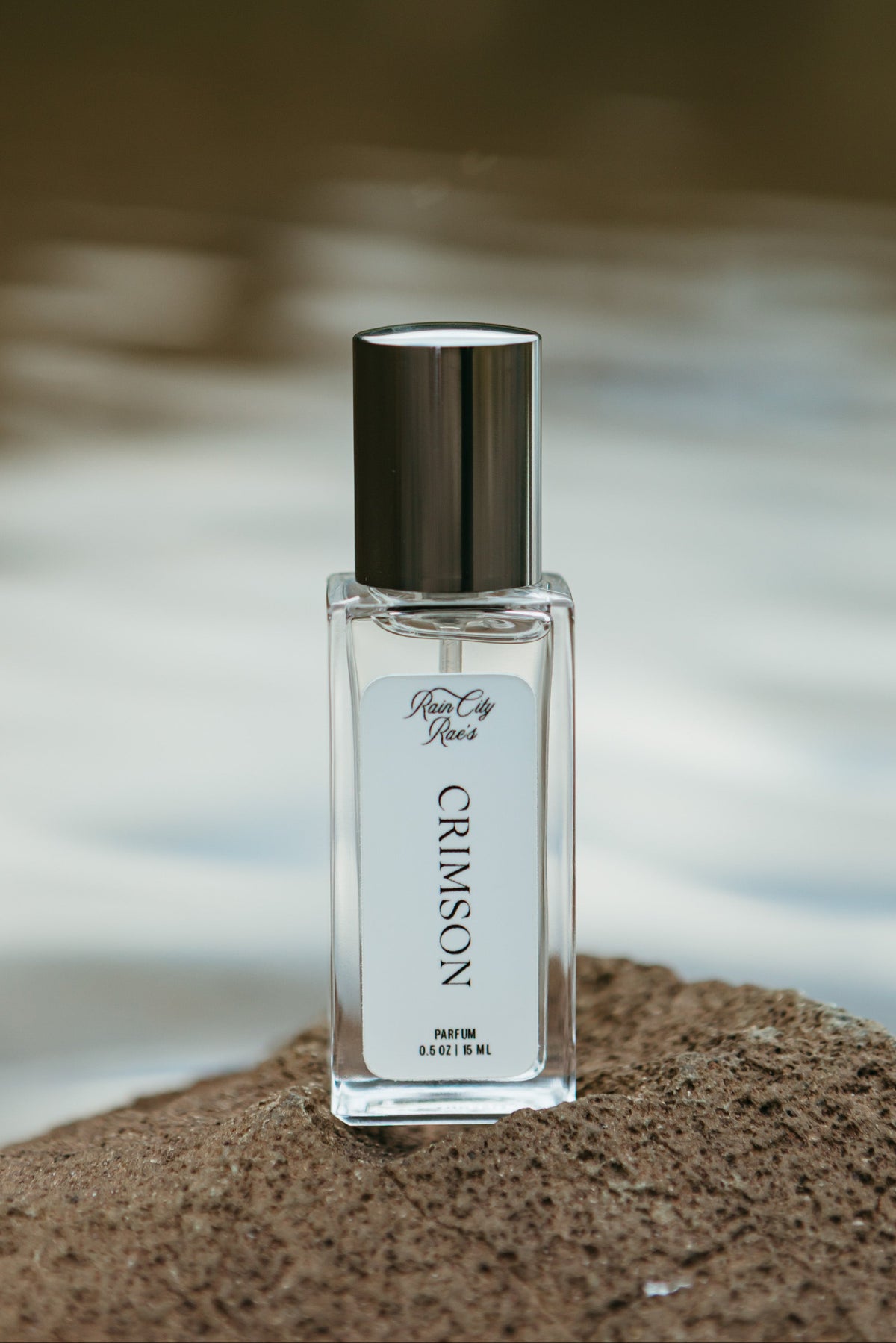 A clear glass perfume bottle labeled 'Crimson' By Rain City Rae's with a grey cap, standing on a textured rock with a blurred outdoor water background.