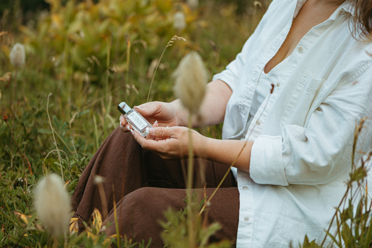 A person sitting outdoors in a grassy field holds a small clear bottle of perfume with a silver cap. The person is wearing a white button-up shirt and brown pants, with blurred green and beige grass in the background. Forever and Always By Rain City Rae's