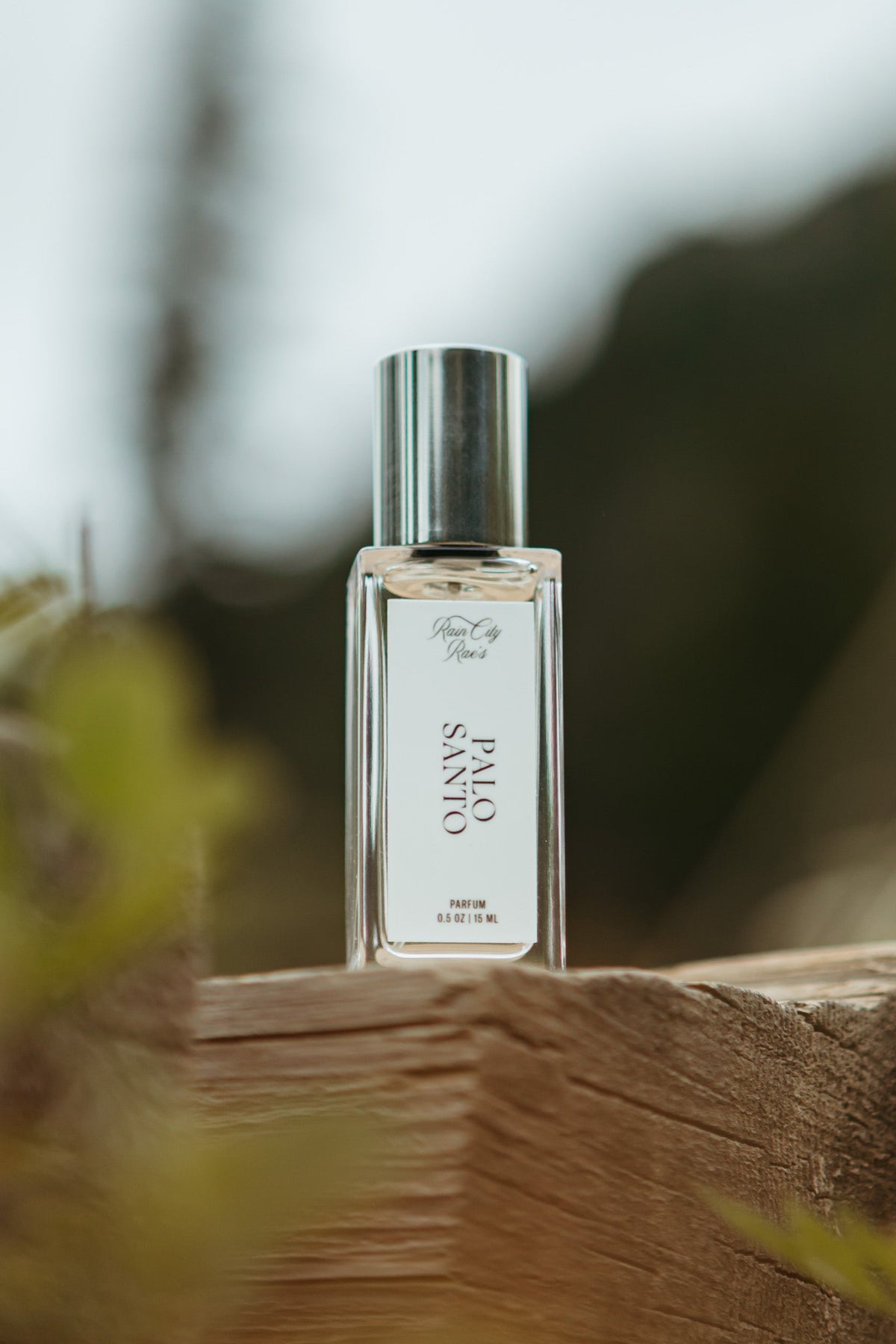 A clear rectangular perfume bottle with a silver cap stands on a rustic wooden surface outdoors, with blurred foliage and hills in the background. Palo Santo By Rain City Rae's