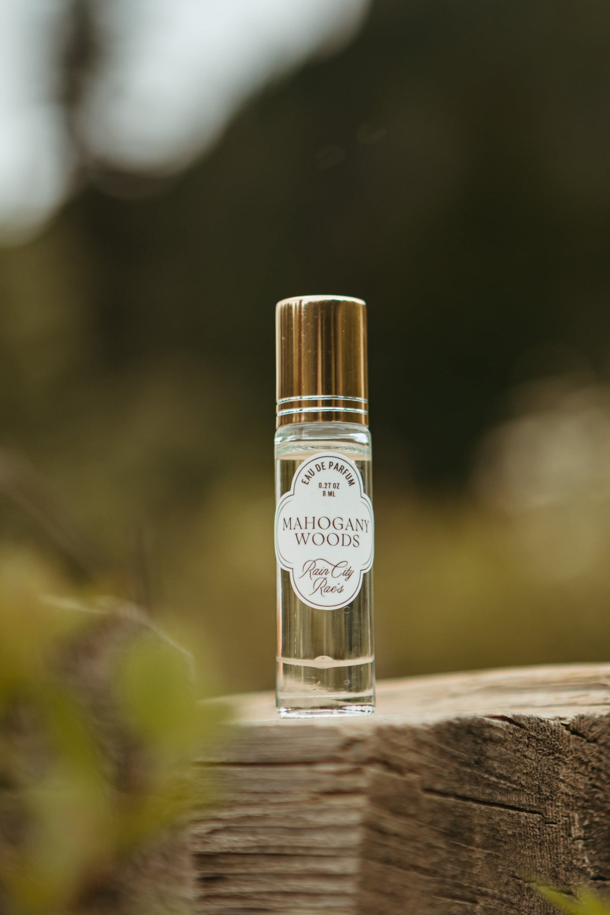 A small clear glass roller bottle perfume with a gold cap and a white label reading 'Mahogany Woods' stands on a weathered wooden log outdoors, with a blurred natural background.