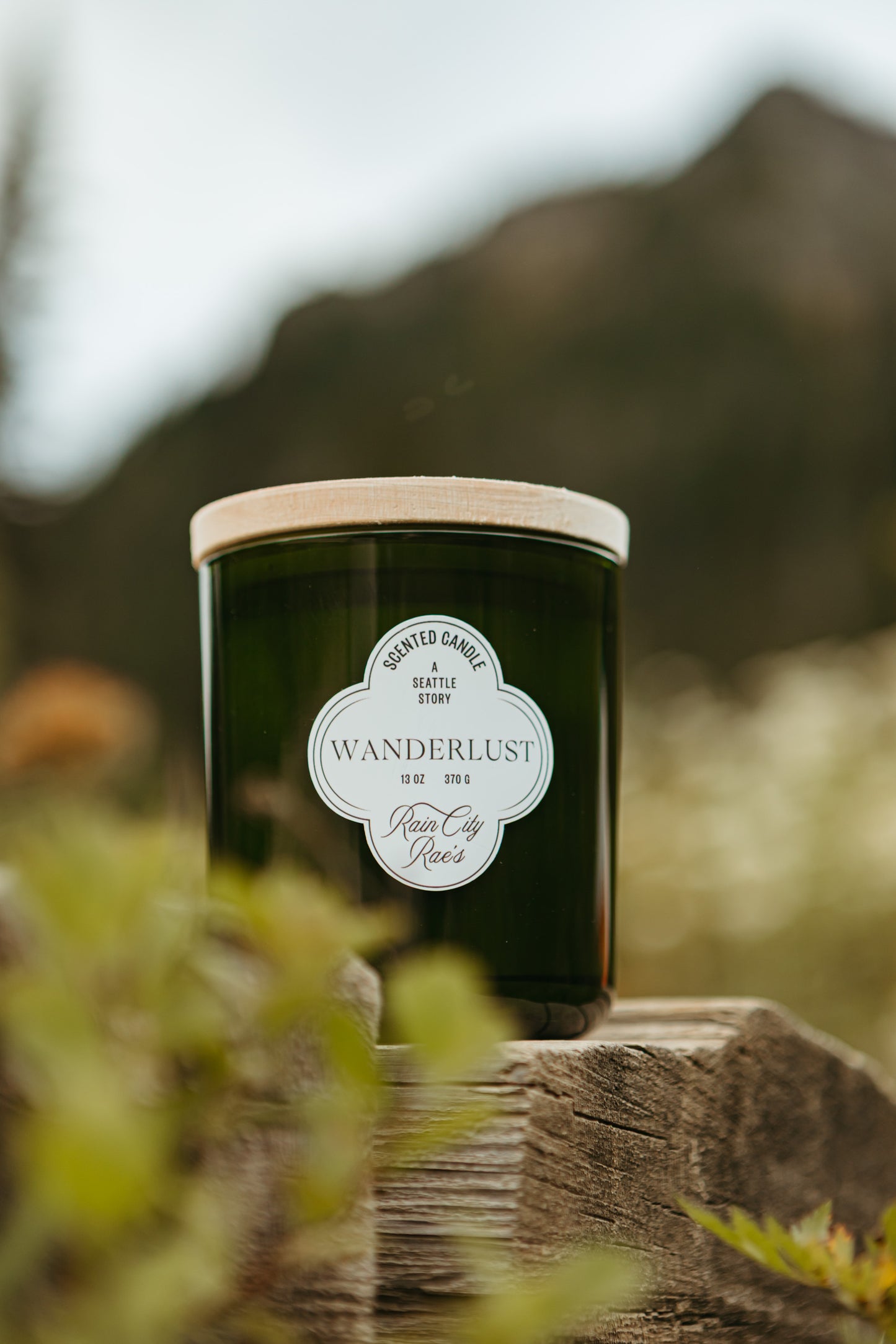A dark green glass jar candle with a light wood lid sits on a rustic wooden surface outdoors, with a blurred mountain and foliage in the background. The front label reads Wanderlust by Rain City Rae's.