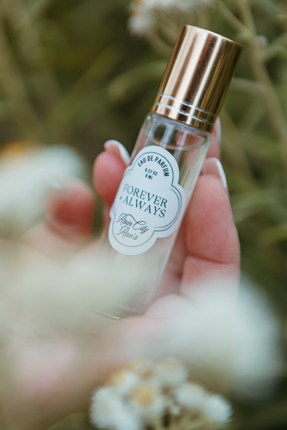 A hand holding a clear glass perfume roller bottle with a shiny gold cap and a white decorative label that reads 'FOREVER and ALWAYS'. The background is a soft-focus outdoor scene with green foliage and white flowers.