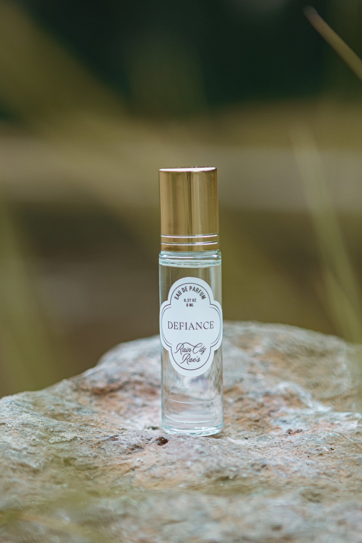 A small clear glass perfume bottle with a metallic gold cap and a white flower label named Dedfiance, standing on a textured stone with a blurred natural background.