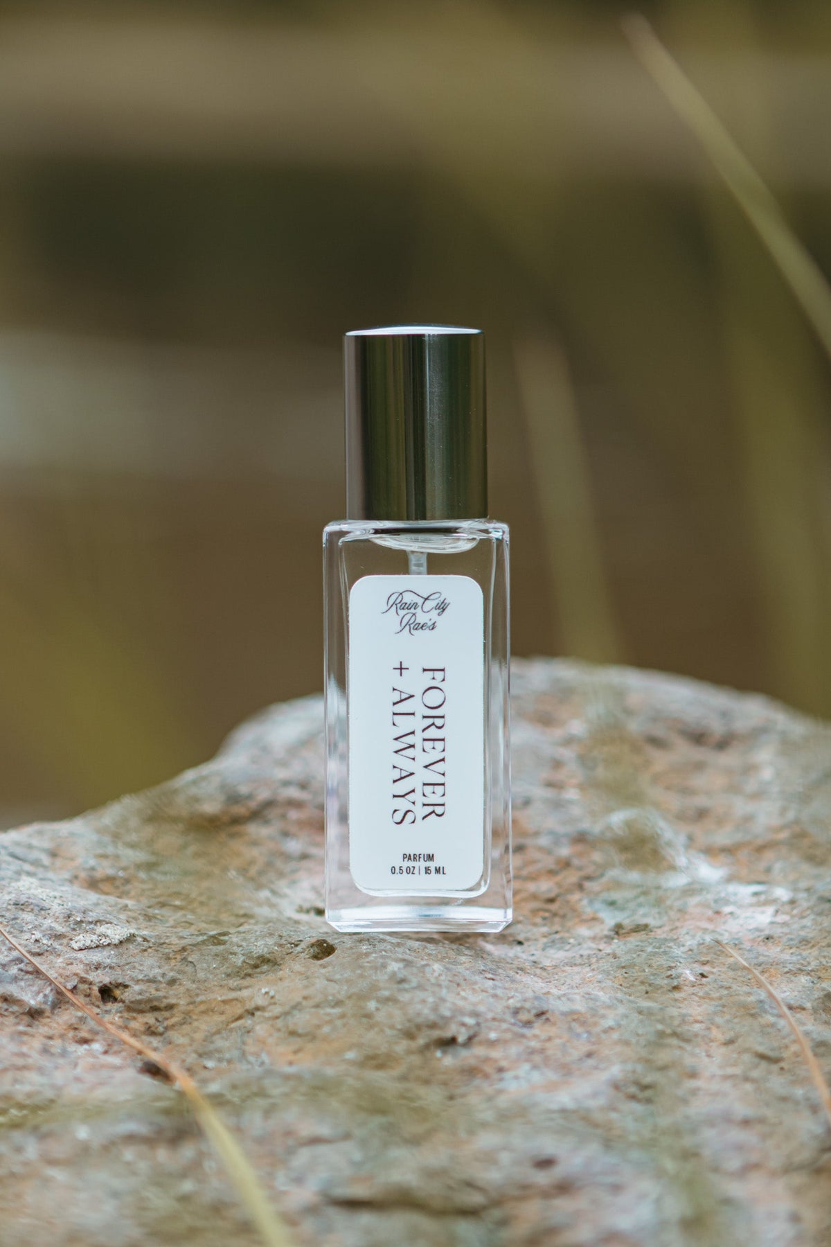 A small clear glass perfume bottle with a silver cap and a white label reading 'Forever and Always' By Rain City Rae's, standing on a textured stone with a blurred natural background.