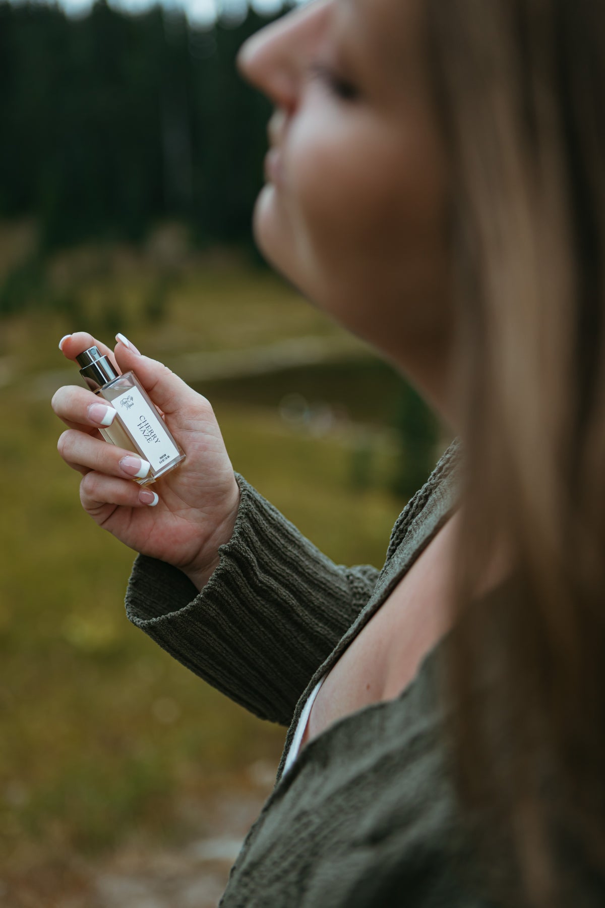 A woman in a ribbed olive green sweater holds a small clear glass perfume bottle with a silver cap, smelling the fragrance outdoors with a blurred forest background. Cherry Haze botanical perfume By Rain City Rae's