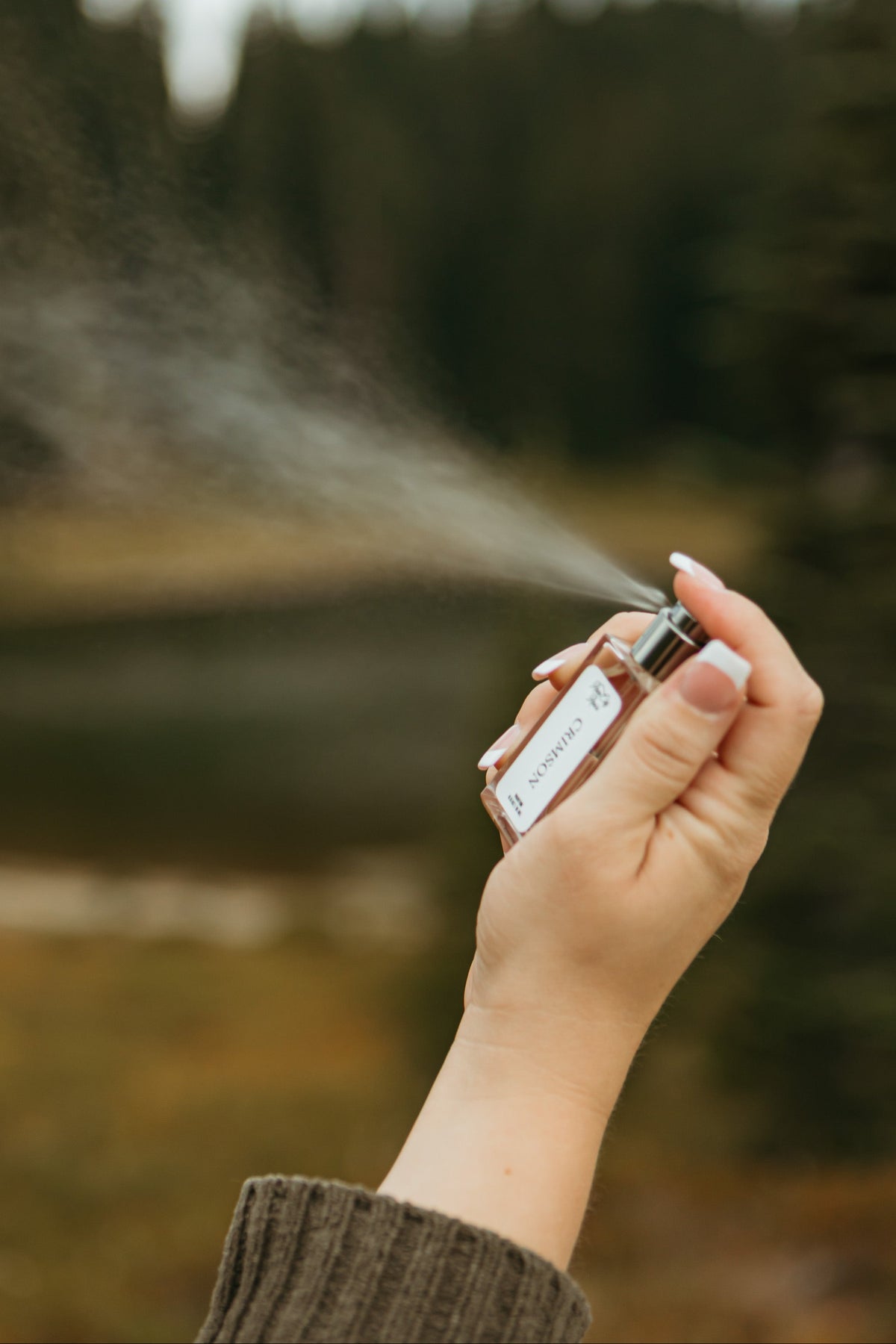 A hand holding a small perfume bottle that is spraying a fine mist, set against a blurred outdoor background of a forest and lake. By Rain City Rae's