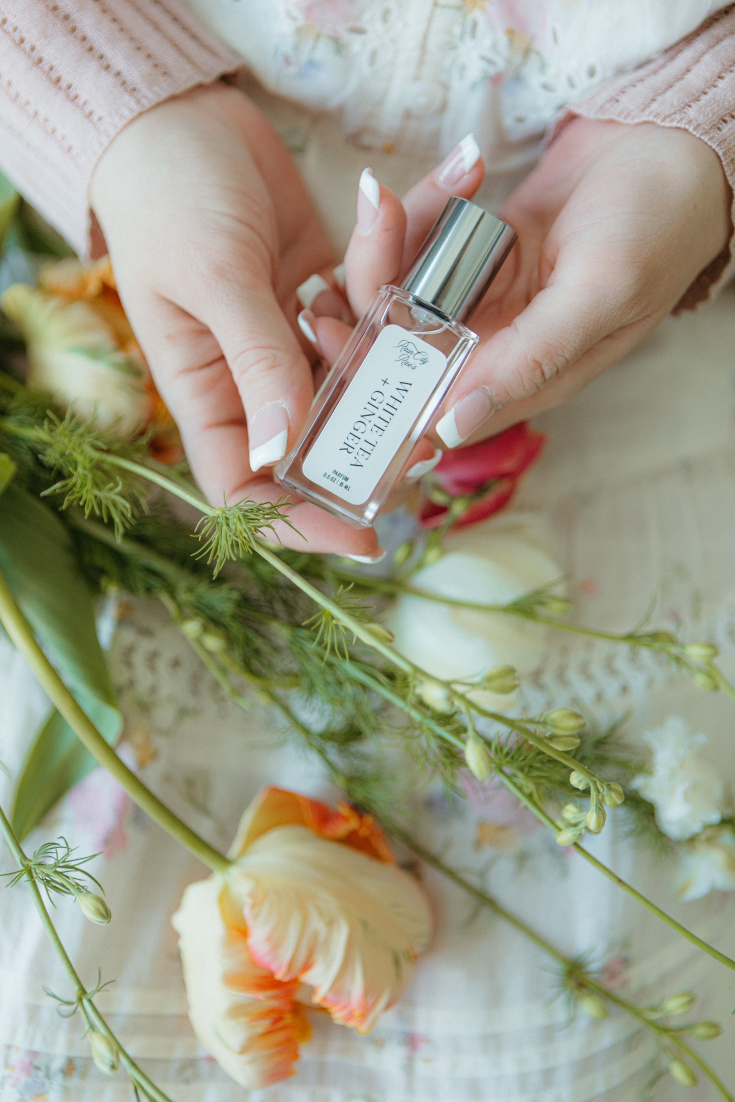 Hands holding a perfume bottle with flowers in the background white tea and ginger scent; pinks, whites, greens, and soft oranges